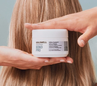 Dwie dłonie trzymają puszkę „GOLDWELL DUALSENSES BOND PRO 60Sec Treatment” przed długimi, blond włosami.